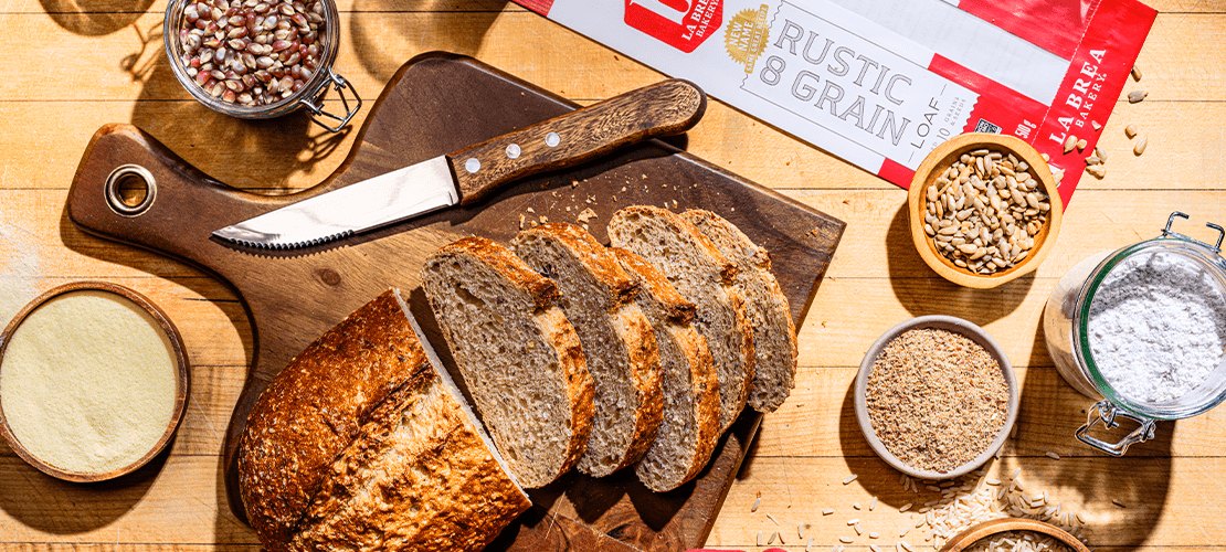 A sliced loaf of Rustic 8 artisan bread on a cutting board with a knife surrounded by raw ingredients used in bread making