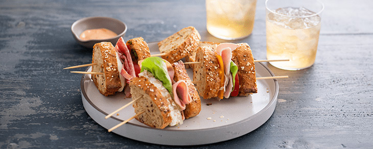 Multi-grain artisan bread sandwich pieces on skewers laying on a white plate with a slate background