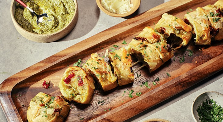 Sliced Sun dried tomato and pesto garlic bread on wood serving board with spices and sauce