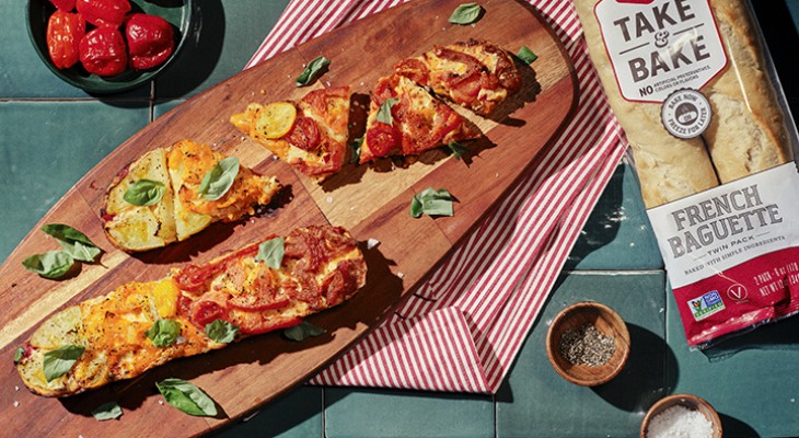 Tomato pie baguette pizza on a wood serving board with a pack of La Brea Take and bake baguette artisan bread