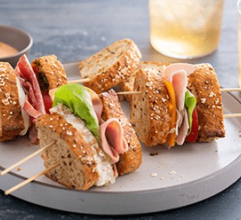 Multi-grain artisan bread sandwich pieces on skewers laying on a white plate with a slate background